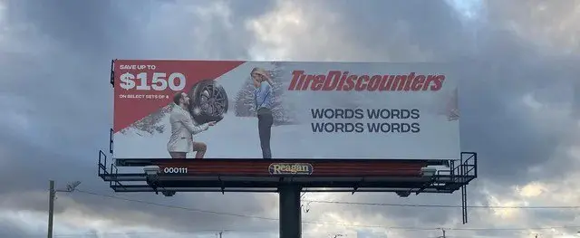 This billboard has a copywriting error. The billboard shows an image of a man proposing to a woman with a tire as a ring, the name "TireDiscount" and then text copywriting that says "WORDS WORDS WORDS WORDS" which was likely supposed to be replaced with marketing copy.